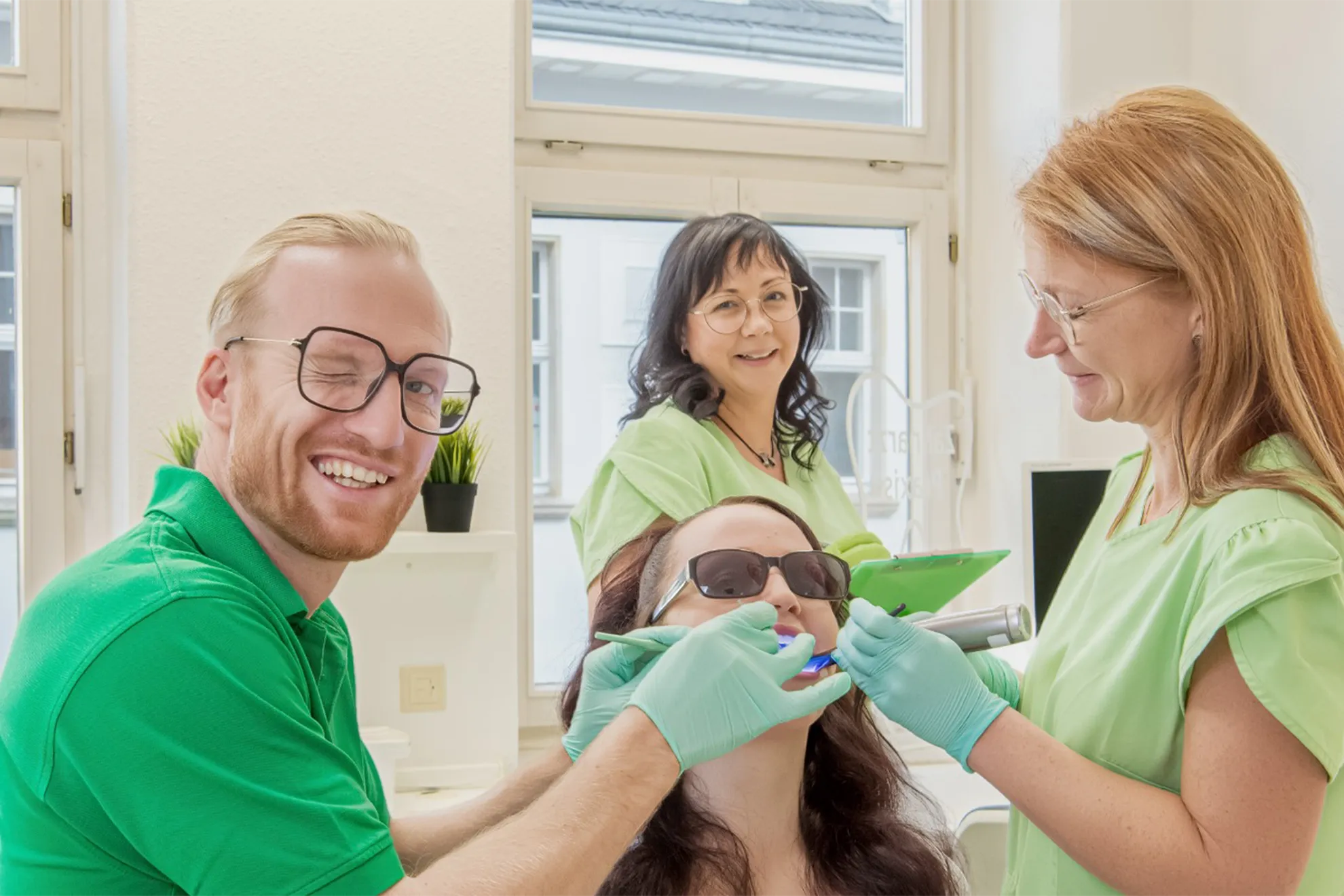 Zahnarzt und 2 ZFA im Behandlungsraum mit einer Patientin, Zahnarzt blinzelt in die Kamera