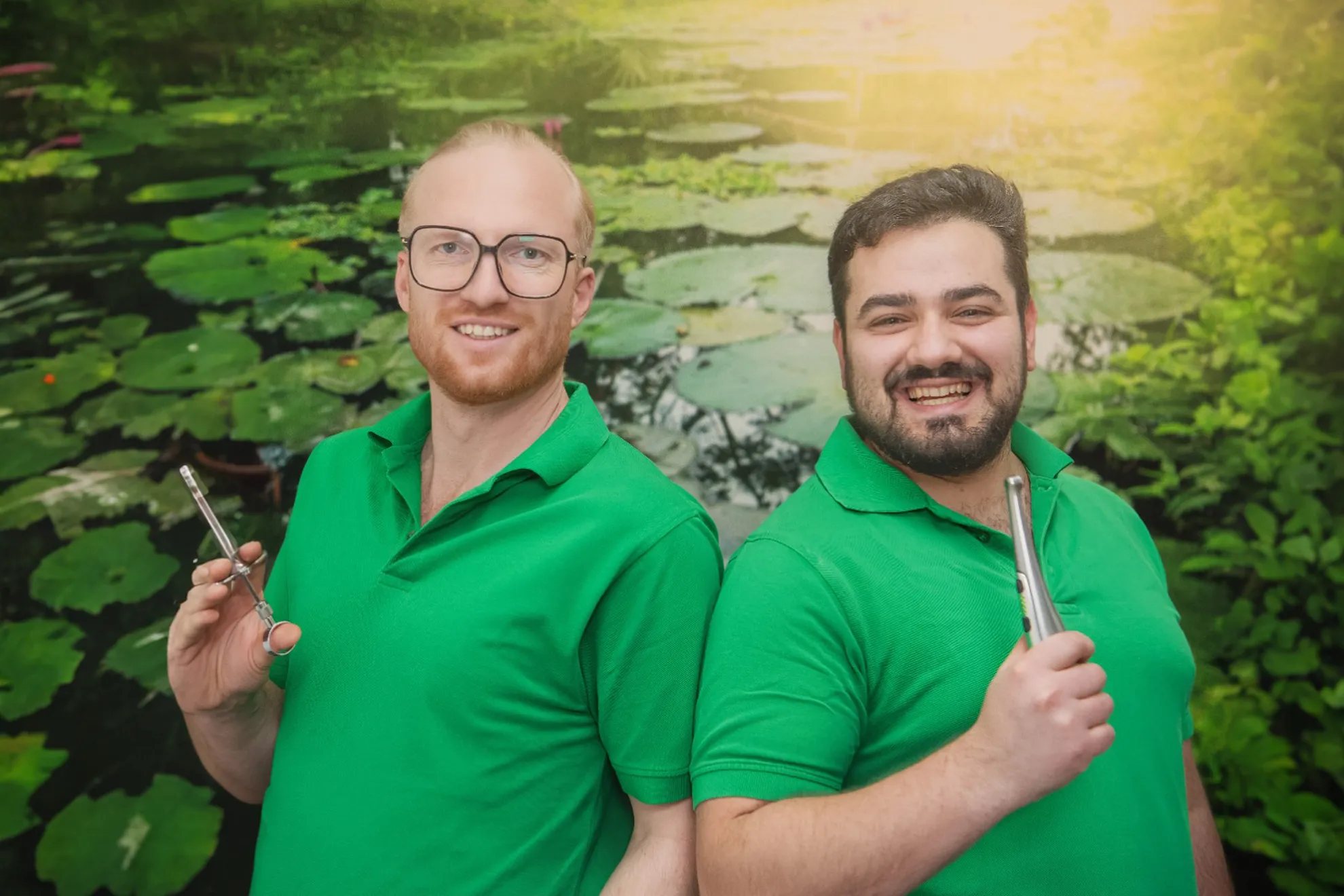 2 Zahnärzte lächeln in die Kamera, Schulter an Schulter mit zahnmedizinsichen Werkzeugen in der Hand